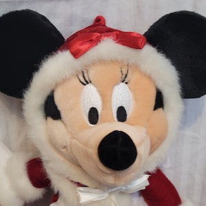 K&ouml;nnte beinhalten: Ein Pl&uuml;schtier von Minnie Mouse, das ein rot-wei&szlig;es Winteroutfit mit einer roten Schleife tr&auml;gt. Sie hat schwarze Ohren und eine schwarze Nase.