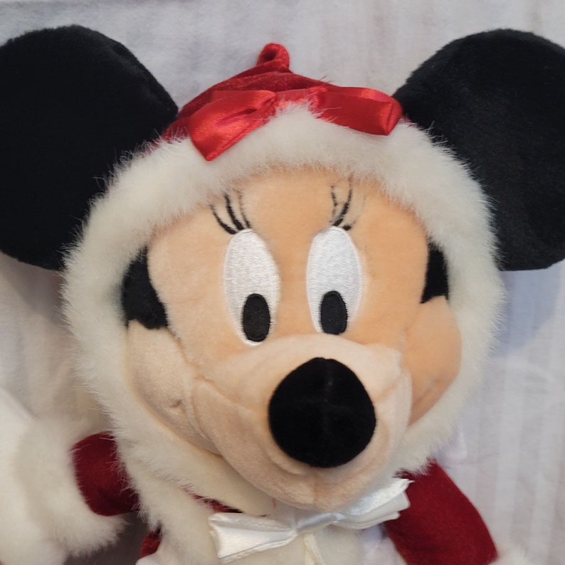 K&ouml;nnte beinhalten: Ein Pl&uuml;schtier von Minnie Mouse, das ein rot-wei&szlig;es Winteroutfit mit einer roten Schleife tr&auml;gt. Sie hat schwarze Ohren und eine schwarze Nase.
