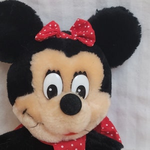 Puede incluir: Un juguete de peluche de Minnie Mouse con orejas negras, un lazo rojo con lunares blancos y un vestido rojo con lunares blancos.
