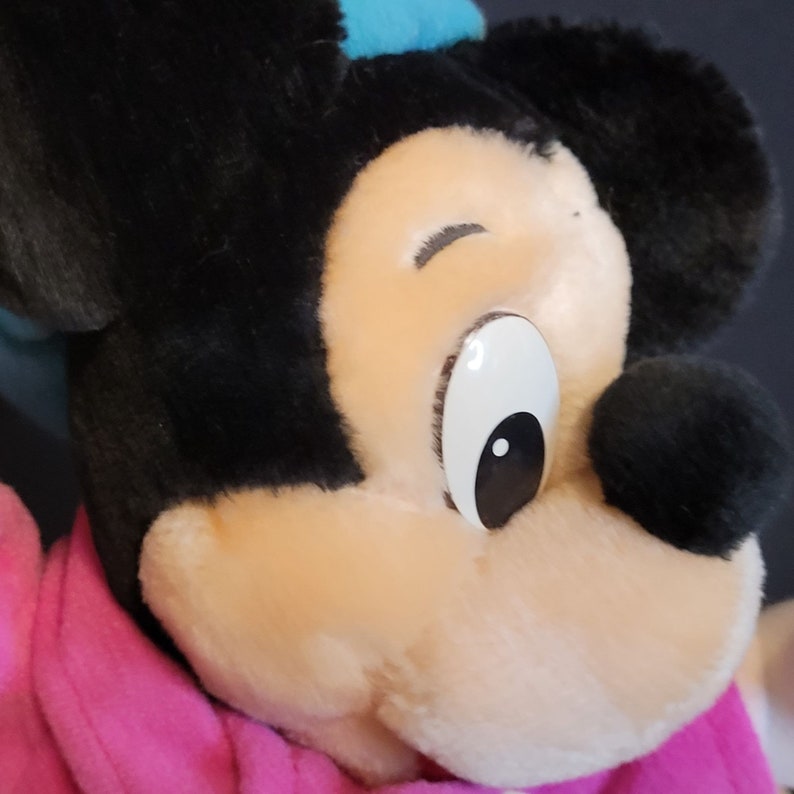 Op de afbeelding: Close-up van een pluche Mickey Mouse speelgoed met zwart haar, een roze sjaal en een groot wit oog met een zwarte pupil.
