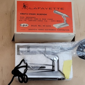 Op de afbeelding: Vintage Lafayette Versatile Dynamic Microphone in de originele doos. De doos is oranje en wit met het Lafayette-logo en productdetails. De microfoon is zilverkleurig en wordt geleverd met een bureaustandaard en snoer.
