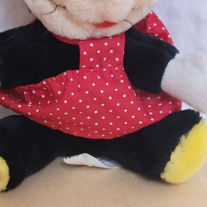 Puede incluir: Un rat&oacute;n de peluche que lleva un vestido rojo con lunares blancos. El rat&oacute;n tiene ojos negros, una nariz negra y una boca roja. El rat&oacute;n est&aacute; sentado sobre una superficie de madera.