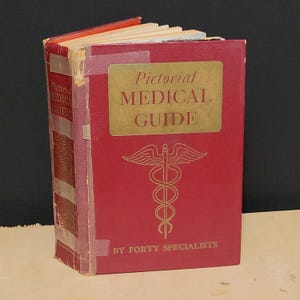 以下が含まれることがあります： 「Pictorial Medical Guide」というタイトルと金色の文字、医療シンボルが特徴のヴィンテージの赤い本。本の背表紙にはタイトルと若干の摩耗が見られます。表紙には「By Forty Specialists」とも書かれています。