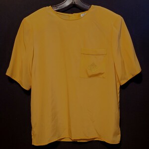 May include: A yellow short-sleeved blouse with a pocket on the left side. The pocket has a small embroidered logo that says "C.C."