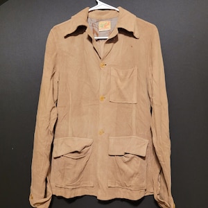 Op de afbeelding: Een bruin corduroy jasje met een kraag, drie knopen en vier voorzakken. De jas hangt aan een witte hanger tegen een donkere achtergrond. Een klein label is zichtbaar bij de kraag.
