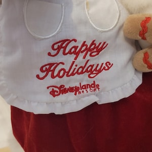 K&ouml;nnte beinhalten: Wei&szlig;er Stoff mit rotem gesticktem Text, der "Frohe Weihnachten" lautet, und einem Logo, das "Disneyland Resort" lautet.