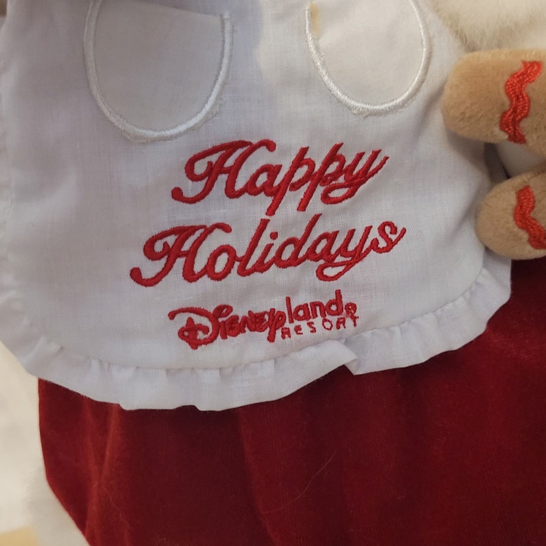 K&ouml;nnte beinhalten: Wei&szlig;er Stoff mit rotem gesticktem Text, der "Frohe Weihnachten" lautet, und einem Logo, das "Disneyland Resort" lautet.
