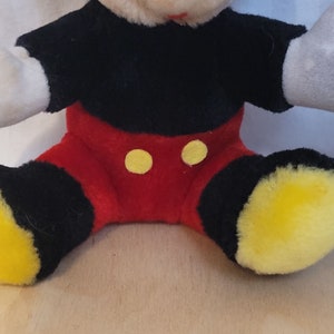 Puede incluir: Un juguete de peluche de Mickey Mouse sentado con las piernas cruzadas. El juguete lleva una camisa negra, pantalones cortos rojos con dos botones amarillos y zapatos amarillos.