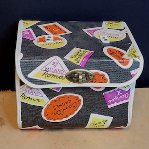 May include: A black and white travel-themed cosmetic case with a white trim. The case has a variety of colorful stickers with city names and landmarks, including "Milano Roma", "Zurich", "Cannes Carlton", "Claridge London", and "Hotel Gra".