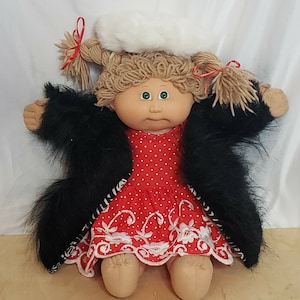 Könnte beinhalten: Eine Cabbage Patch Kid Puppe, die ein rot-weiß gepunktetes Kleid mit weißen Stickereien, einen schwarzen Pelzmantel und einen weißen Hut mit rotem Besatz trägt.