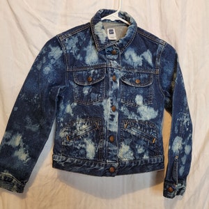 May include: A blue denim jacket with a light blue, bleached pattern. The jacket has four front pockets, button closures, and a collar. The Gap brand label is visible on the inside collar. The jacket is hanging on a white hanger.