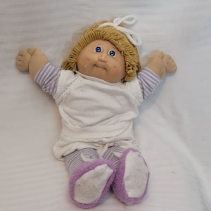Puede incluir: Una muñeca Cabbage Patch Kid con cabello rubio, ojos azules y mejillas sonrosadas. La muñeca lleva una camisa blanca con mangas y pantalones a rayas de color morado y blanco. La muñeca también lleva pantuflas a rayas de color morado y blanco.