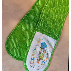 May include: A pair of green oven mitts with white polka dots. One mitt has a white patch with a colorful illustration of a girl in a dress and the text "Love's a starrin".