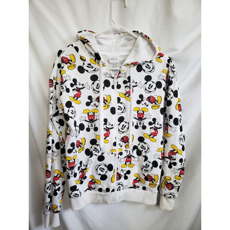 May include: White hooded sweatshirt with a full-coverage print of Mickey Mouse in various poses. The print features black outlines, red shorts, yellow shoes, and black ears. The zipper is white.