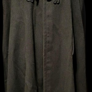 May include: A black cape featuring a fold-down collar and a single decorative black cord frog closure at the neck. The garment has a long, flowing silhouette with a smooth texture, extending downward from the shoulders in a structured, solid design.