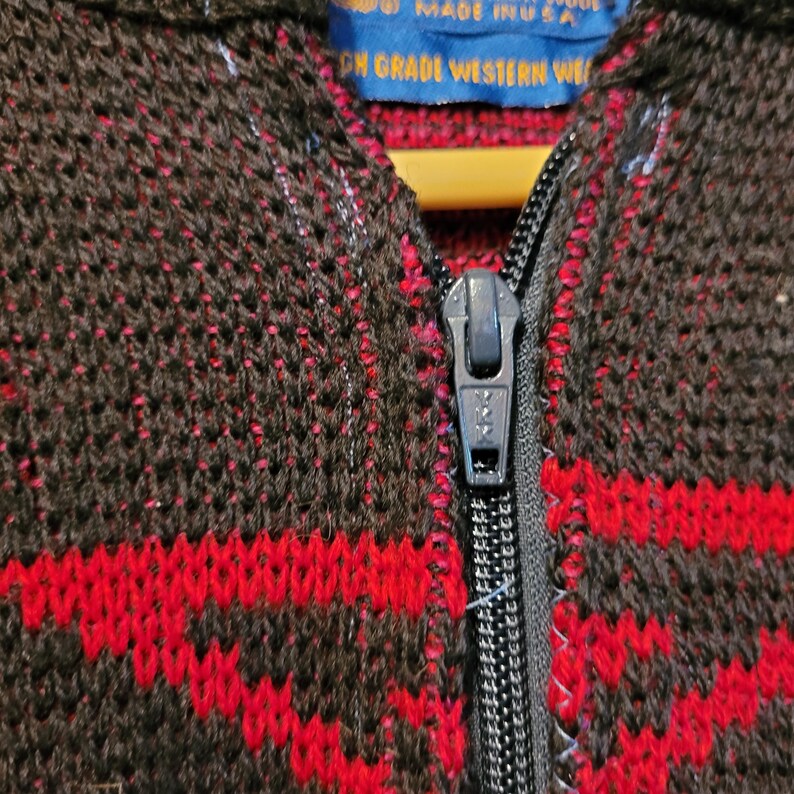 K&ouml;nnte beinhalten: Nahaufnahme eines braunen und roten Strick-Rei&szlig;verschluss-Sweaters. Der Sweater hat ein geometrisches Muster in Rot auf braunem Hintergrund. Ein schwarzer Rei&szlig;verschluss verl&auml;uft in der Mitte. Das Etikett lautet "MADE IN USA".