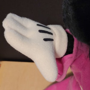 Op de afbeelding: Een pluche speelgoed met een roze en witte outfit. Het speelgoed heeft een witte handschoen met zwarte stiksels op de hand.