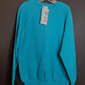 May include: A turquoise blue sweatshirt with a crew neck and raglan sleeves. The sweatshirt has a white tag with the text "Sturdy Sweats" on it.