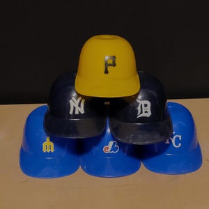 Op de afbeelding: Een verzameling honkbalhelmen in verschillende kleuren. De helmen zijn blauw, geel en marineblauw. Elke helm heeft een teamlogo op de voorkant. De logo's zijn onder meer een "P", "NY", "D", "M", "B" en "C".