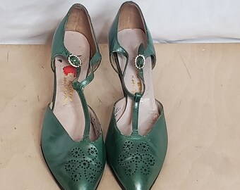 Op de afbeelding: Een paar groene leren hakschoenen met een T-band en een gesp met steentjes. De schoenen hebben een spitse neus en zijn versierd met een bloemmotief van strass.