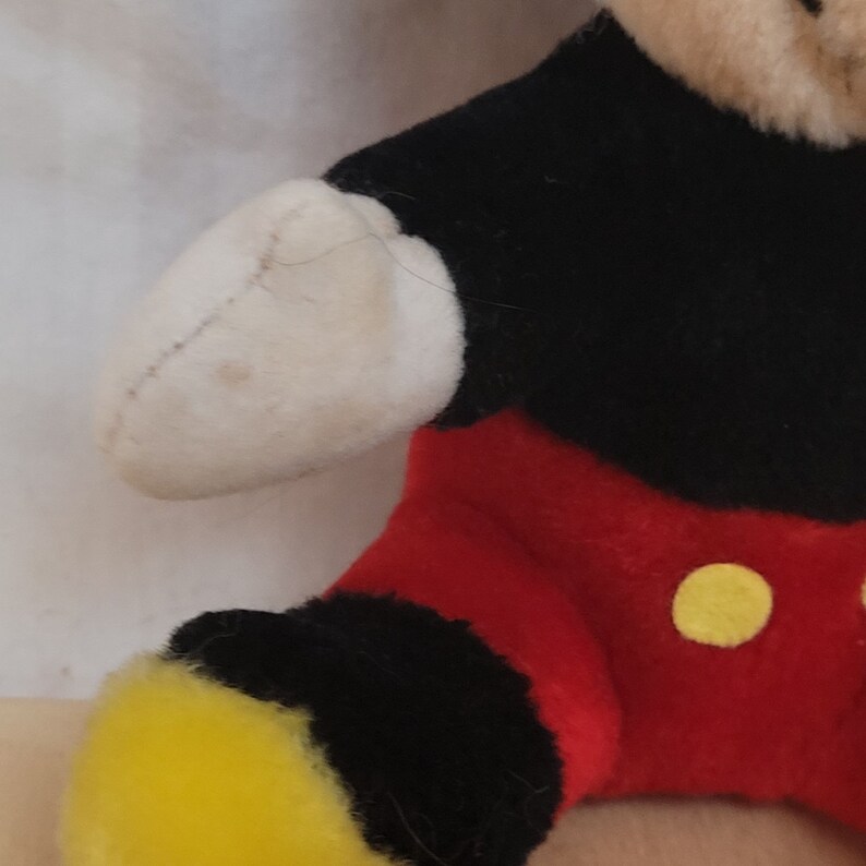 Puede incluir: Un juguete de peluche de Mickey Mouse con ropa negra y roja y botones amarillos. El juguete tiene un brazo blanco con una mancha marr&oacute;n.