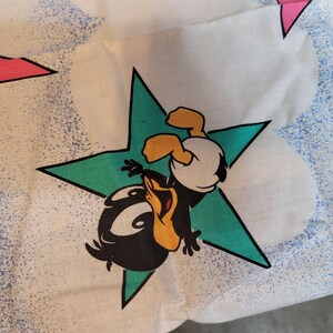 May include: A white fabric with a green star and a black and yellow cartoon duckling wearing a white diaper. The duckling is upside down and has its wings spread out. The fabric has a blue and white speckled pattern.