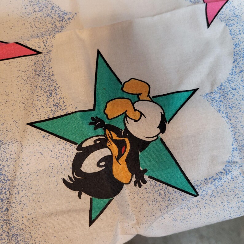May include: A white fabric with a green star and a black and yellow cartoon duckling wearing a white diaper. The duckling is upside down and has its wings spread out. The fabric has a blue and white speckled pattern.