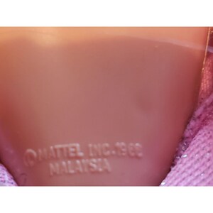 Puede incluir: Primer plano de la cabeza de una mu&ntilde;eca con tono de piel claro. El texto "MATTEL INC. 1966 MALAYSIA" est&aacute; grabado en la cabeza de la mu&ntilde;eca. Se ve una tela rosa y brillante en el lado derecho de la imagen.