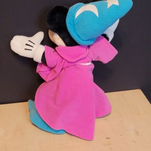 Op de afbeelding: Een pluche Mickey Mouse verkleed als tovenaar. Hij draagt een roze robe, een blauwe hoed met een witte ster en maan, en witte handschoenen.