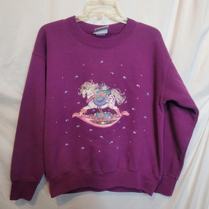 May include: Purple sweatshirt with a white rocking horse with a floral design and a girl riding it. The horse is surrounded by small pink hearts and purple flowers.