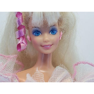 Puede incluir: Primer plano del rostro de una mu&ntilde;eca con ojos azules, labios rosados y cabello rubio. La mu&ntilde;eca tiene pendientes rosas y una cinta rosa en el pelo. Lleva un vestido rosa y blanco.