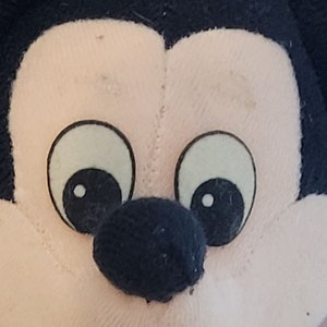 Peut inclure: Gros plan sur un visage en peluche de Mickey Mouse avec des oreilles noires, un nez noir et de grands yeux blancs avec des pupilles noires.
