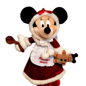 K&ouml;nnte beinhalten: Ein Pl&uuml;sch-Minnie Mouse, gekleidet in ein rotes Samtkleid mit wei&szlig;em Pelzbesatz, einer wei&szlig;en Sch&uuml;rze mit dem Text "Holidays at Disneyland" und einem wei&szlig;en, pelzbesetzten Hut. Sie h&auml;lt einen Holzl&ouml;ffel in der einen Hand und einen Lebkuchenmann in der anderen.