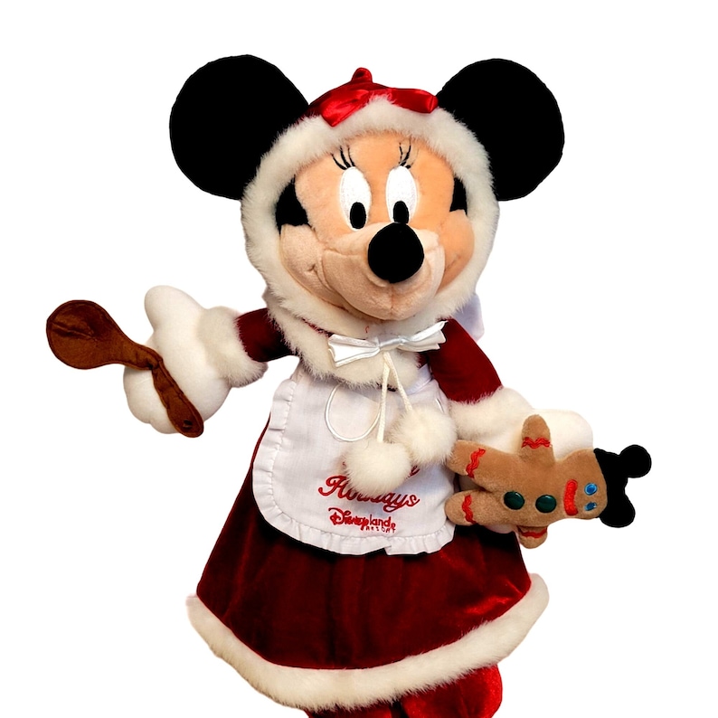 K&ouml;nnte beinhalten: Ein Pl&uuml;sch-Minnie Mouse, gekleidet in ein rotes Samtkleid mit wei&szlig;em Pelzbesatz, einer wei&szlig;en Sch&uuml;rze mit dem Text "Holidays at Disneyland" und einem wei&szlig;en, pelzbesetzten Hut. Sie h&auml;lt einen Holzl&ouml;ffel in der einen Hand und einen Lebkuchenmann in der anderen.
