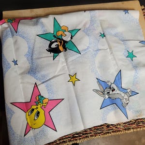 May include: White fabric with a repeating pattern of stars and cartoon characters. The characters are Tweety Bird, Bugs Bunny, and Daffy Duck. The stars are in various colors, including pink, blue, yellow, and green.