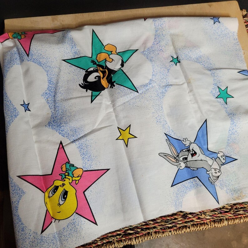 May include: White fabric with a repeating pattern of stars and cartoon characters. The characters are Tweety Bird, Bugs Bunny, and Daffy Duck. The stars are in various colors, including pink, blue, yellow, and green.