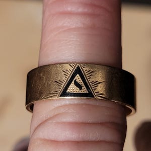 Op de afbeelding: Een goudkleurige ring met een zwart driehoekontwerp. De driehoek bevat een gestileerde 'S'-vorm. De ring heeft geëtste lijnen die uitstralen van de driehoek. De ring wordt aan een vinger gedragen.