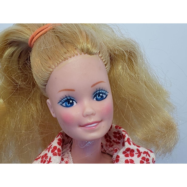 May include: A vintage doll with blonde hair in pigtails, held by an orange elastic band. The doll has blue eyes, pink cheeks, and a smiling face. She wears a white top with a red floral pattern.