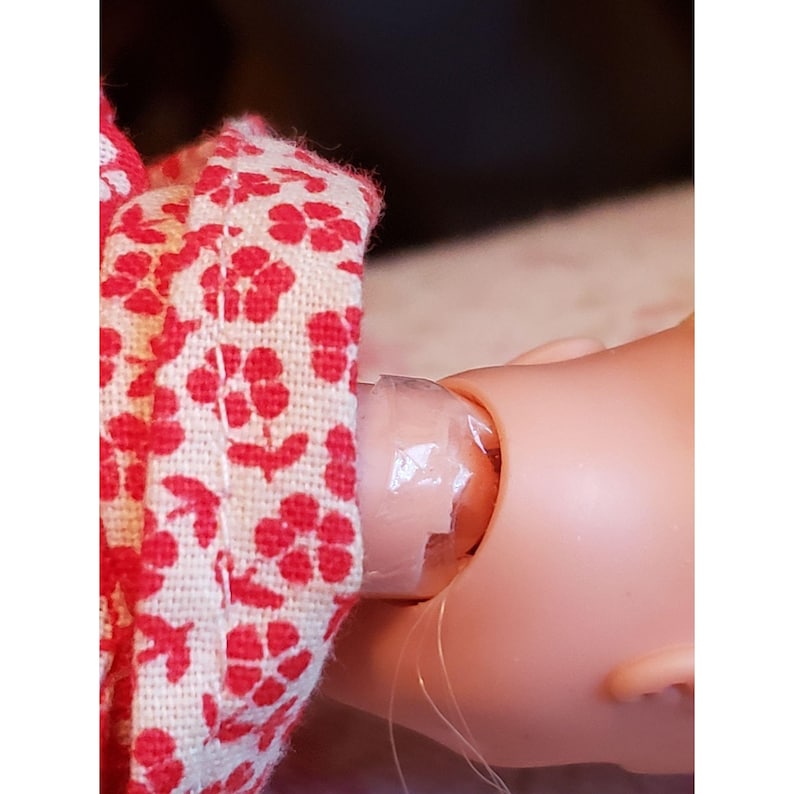 May include: A doll's head and neck are shown, with red floral fabric attached to the neck. The doll's neck is wrapped in clear tape. The fabric is white with red flowers.