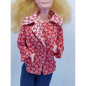 May include: A red jacket with a white floral pattern, featuring a wide collar and white lining. The jacket is buttoned. The doll is also wearing blue denim jeans.