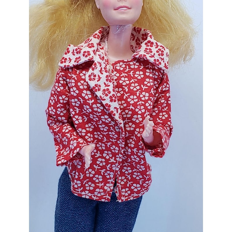May include: A red jacket with a white floral pattern, featuring a wide collar and white lining. The jacket is buttoned. The doll is also wearing blue denim jeans.