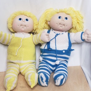 May include: Two Cabbage Patch Kids dolls wearing knitted outfits. One doll is wearing a yellow and white striped outfit and the other is wearing a blue and white striped outfit.