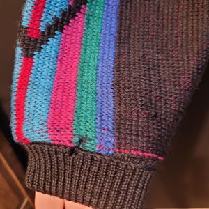 K&ouml;nnte beinhalten: Nahaufnahme eines dunkelgrauen Pullover&auml;rmels mit geripptem B&uuml;ndchen. Der &Auml;rmel hat ein farbenfrohes Streifenmuster in Rot, Blau, Gr&uuml;n und Pink. Das B&uuml;ndchen ist einfarbig dunkelgrau.
