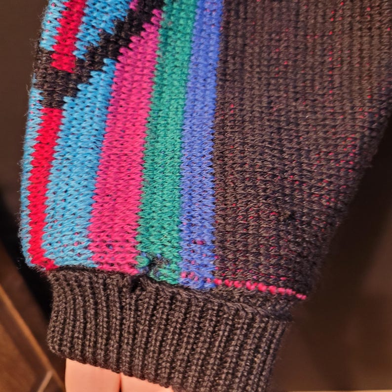K&ouml;nnte beinhalten: Nahaufnahme eines dunkelgrauen Pullover&auml;rmels mit geripptem B&uuml;ndchen. Der &Auml;rmel hat ein farbenfrohes Streifenmuster in Rot, Blau, Gr&uuml;n und Pink. Das B&uuml;ndchen ist einfarbig dunkelgrau.