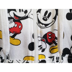 May include: White zippered hoodie with a repeating pattern of Mickey Mouse in various poses. The hoodie has a white zipper and white drawstrings.