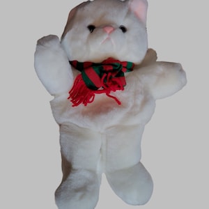 Könnte beinhalten: Weißer Plüschkatze mit einem rot-grünen gestreiften Schal mit roter Quaste. Die Katze steht auf ihren Hinterbeinen mit erhobenen Vorderpfoten.