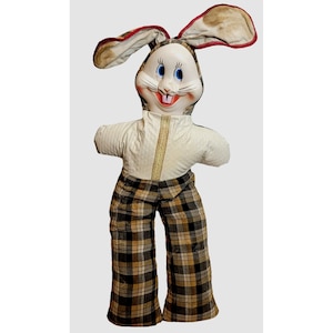 Può includere: Un giocattolo di coniglio di peluche vintage con una faccia bianca, occhi blu e una bocca rossa. Il coniglio indossa una camicia bianca e pantaloni a quadri marroni e neri.