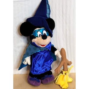 K&ouml;nnte beinhalten: Ein Pl&uuml;sch-Minnie Mouse, verkleidet als Hexe f&uuml;r Halloween. Sie tr&auml;gt einen blauen und t&uuml;rkisfarbenen Hexenhut, ein blaues Kleid mit einem t&uuml;rkisfarbenen, gepunkteten Umhang und lila Schuhe mit Fledermausfl&uuml;geln. Sie h&auml;lt einen braunen und gelben Besen.