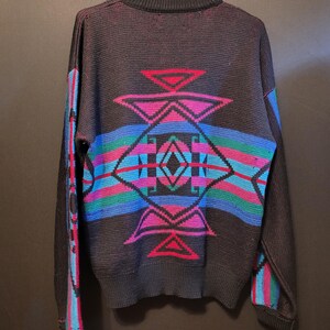 K&ouml;nnte beinhalten: Schwarzer Strickpullover mit geometrischem Muster in Rot, Pink, Blau und Gr&uuml;n. Der Pullover hat einen Rundhalsausschnitt und lange &Auml;rmel mit Streifen. Die R&uuml;ckseite zeigt ein gro&szlig;es, farbenfrohes Muster.