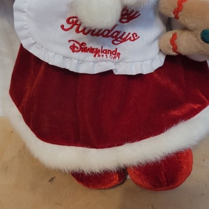 K&ouml;nnte beinhalten: Ein rotes und wei&szlig;es Pl&uuml;schtier, das als Lebkuchenfrau verkleidet ist. Das Spielzeug tr&auml;gt eine wei&szlig;e Sch&uuml;rze mit dem Text "Happy Holidays Disneyland Resort".